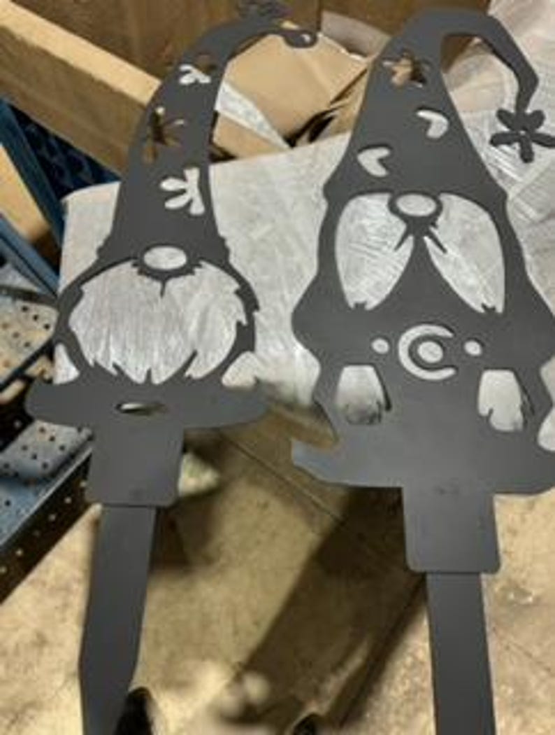 May include: Two black metal garden gnomes with a snowflake design. The gnomes are standing upright and have a whimsical design.