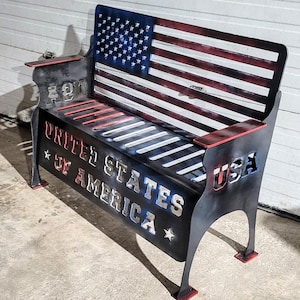 Może przedstawiać: Metalowa ławka z motywem flagi amerykańskiej. Ławka ma wycięty napis "UNITED STATES OF AMERICA" i "USA". Czerwone akcenty na podłokietnikach i nóżkach. Idealna do użytku na zewnątrz.