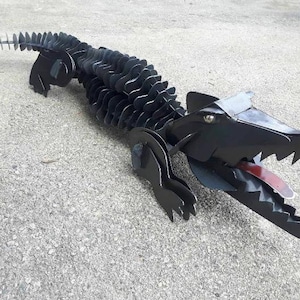 Peut inclure: Une sculpture d'alligator en métal noir avec une langue rouge. L'alligator est fait de pièces de métal superposées, ce qui lui donne un aspect texturé. La sculpture est posée sur une surface en béton grise.