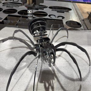 Peut inclure: Sculpture d'araignée en métal sombre avec huit longues pattes incurvées et un corps segmenté. Le corps de l'araignée est fait de pièces de métal superposées et projette une ombre sur la surface en dessous. La sculpture est probablement faite à la main.