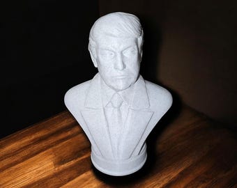 May include: A white bust sculpture of a man with short, dark hair. The bust is wearing a suit jacket and tie. The sculpture is on a wooden surface.