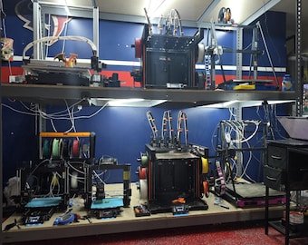 May include: A workshop featuring multiple 3D printers on shelving units. The printers are black and orange, with various spools of filament in different colors. The background is a blue wall with red accents, and the floor is red.
