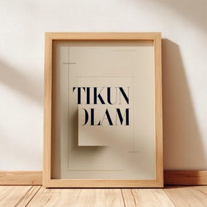 May include: A framed print with the words "TIKUN OLAM" in navy blue, set against a beige background. The print is in a light brown wooden frame, leaning against a white wall and wooden floor.