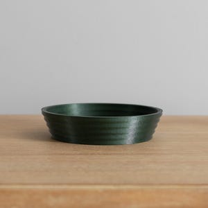 May include: A dark green, round, ceramic bowl with a slightly rippled texture. The bowl is sitting on a light brown wooden surface.
