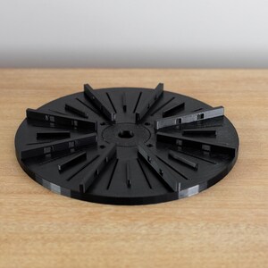 May include: A black plastic disc with multiple radial slots, resembling a sunburst design. The disc has a central hole and is designed for use with a turntable.