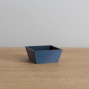 May include: A small, square, blue tray made of cardboard. The tray is sitting on a light brown wooden surface.