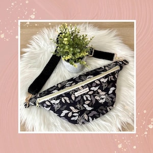 Puede incluir: Riñonera negra con cremallera dorada y correa negra ajustable. La riñonera tiene un estampado de hojas blancas y doradas.