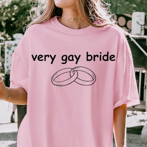 Puede incluir: Camiseta rosa claro con la frase "very gay bride" y dos anillos entrelazados en negro. La camiseta es holgada y parece estar hecha de un material suave. El diseño es sencillo.