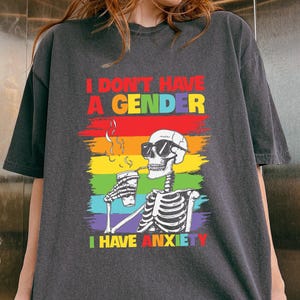 Può includere: T-shirt grigio scuro con la scritta "I DON'T HAVE A GENDER I HAVE ANXIETY" sopra una grafica di uno scheletro con occhiali da sole e una tazza. La grafica include un design arcobaleno. La maglietta è di vestibilità ampia.