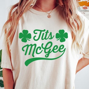 May include: Cream-colored t-shirt with the text "Tits McGee" in green, accompanied by green shamrock graphics. The shirt is a casual style, suitable for St. Patrick's Day or a fun, playful look.