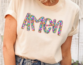 Puede incluir: Una camiseta blanca con la palabra "Amén" escrita en letras florales de colores. La camiseta la lleva puesta una persona con pantalones vaqueros azules.