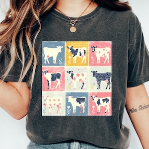 以下が含まれることがあります： 暗いグレーのTシャツに、9匹のカラフルな牛がグリッド状に描かれたグラフィックがプリントされています。牛は白地に黒の斑点、黒地に白の斑点、黒と白の牛です。グラフィックの背景は、黄色、ピンク、青、緑が混ざり合った色です。