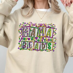 May include: Beige sweatshirt with the text "Does your Mama know how you got those beads?" in a colorful, blocky design. The text is surrounded by green, purple, and yellow beads, evoking a festive atmosphere. The sweatshirt is a casual, comfortable style.