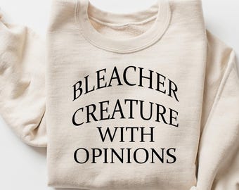 Puede incluir: Una sudadera de color crema con la frase "BLEACHER CREATURE WITH OPINIONS" impresa en negro. Las mangas están enrolladas y la sudadera está colocada sobre una superficie blanca. Una prenda de vestir informal y cómoda.