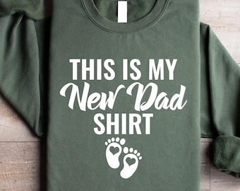 May include: Olive green sweatshirt with the text "THIS IS MY New Dad SHIRT" in white script. Below the text are two white baby footprints with heart-shaped details. The sweatshirt is folded on a wooden surface.