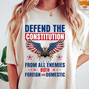 May include: A cream-coloured t-shirt featuring the text "DEFEND THE CONSTITUTION" above an eagle with wings designed like the American flag. The shirt also reads "FROM ALL ENEMIES BOTH FOREIGN AND DOMESTIC".