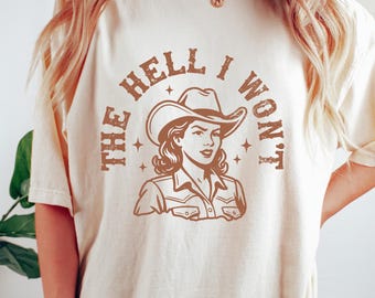 May include: Cream-colored t-shirt featuring a brown graphic of a cowgirl with the text "THE HELL I WON'T." The design includes a cowgirl wearing a hat. The shirt is a casual wear item.