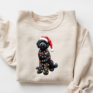 May include: A cream-coloured sweatshirt with a graphic of a black dog wearing a Santa hat and wrapped in Christmas lights. The dog is the central focus, with the lights and hat adding festive colour.
