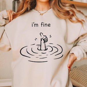 Może przedstawiać: Biała bluza z napisem "i'm fine" i czarną ilustracją dłoni pokazującej kciuk w górę, podczas gdy jest zanurzona w wodzie.