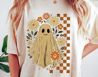 May include: A cream-coloured t-shirt with a smiling ghost design surrounded by daisies and leaves. A checkered pattern is on the right side of the ghost. The shirt has short sleeves and a relaxed fit, suitable for casual wear.