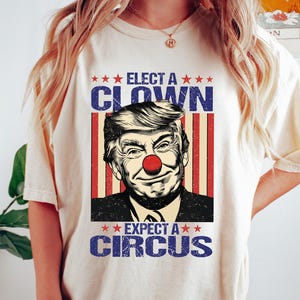 Può includere: T-shirt color crema con grafica di una persona con naso da clown. Il testo recita "ELECT A CLOWN EXPECT A CIRCUS" in blu e rosso. La maglietta è indossata.