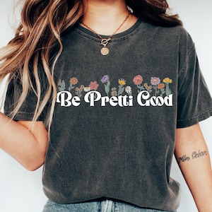 May include: Dark gray t-shirt with the text "Be Pretti Good" in white, above a row of colorful flowers. The shirt is paired with blue jeans and white slip-on shoes. The overall style is casual and relaxed.