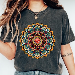 May include: Dark gray t-shirt featuring a vibrant, colorful mandala design. The mandala is a circular pattern with shades of red, yellow, blue, and green. The shirt is paired with blue jeans and white sneakers. The image also includes a necklace and bracelets.