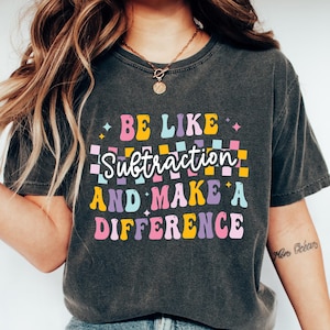 May include: Dark gray t-shirt with the text "BE LIKE Subtraction AND MAKE A DIFFERENCE" in colorful, retro-style lettering. The shirt is paired with blue jeans and white sneakers. The overall aesthetic is casual and comfortable.