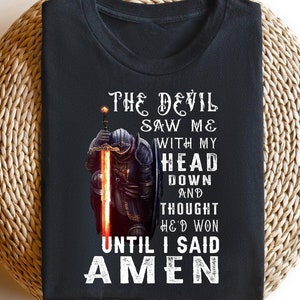 Puede incluir: Camiseta negra con un gráfico blanco de un caballero sosteniendo una espada. El texto en el gráfico dice "The Devil Saw Me With My Head Down And Thought He'd Won Until I Said Amen".