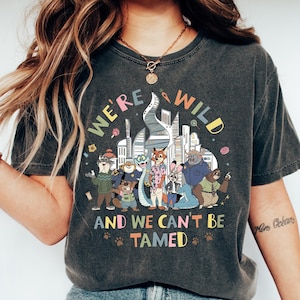 May include: Dark gray t-shirt with the text "WE'RE WILD AND WE CAN'T BE TAMED" above a colorful cartoon illustration of animal characters in a city setting. The shirt is worn with blue jeans and white sneakers.