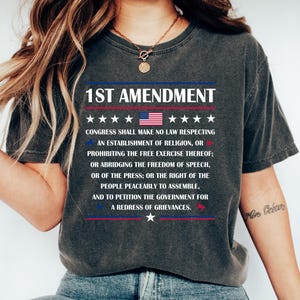May include: Dark grey t-shirt with the text "1ST AMENDMENT" and the US flag graphic. The shirt displays the amendment's text. The shirt is worn with blue jeans and white trainers. The design includes stars and stripes.