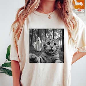 May include: Cream-colored t-shirt featuring a black and white image of a cat taking a selfie with a ghost in a forest. The cat has wide eyes and an open mouth. The shirt is a casual style.