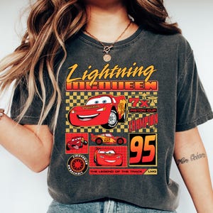 May include: Dark grey t-shirt with a Lightning McQueen design from the Cars film. The graphic includes the text "Lightning McQueen", "95", and "Piston Cup Champion", alongside images of the character. A casual, comfortable top.