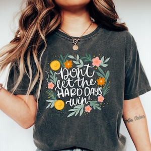 May include: Dark gray t-shirt with the text "Don't let the HARD DAYS win!" surrounded by a floral design with orange and pink flowers. The shirt is paired with blue jeans and white shoes.