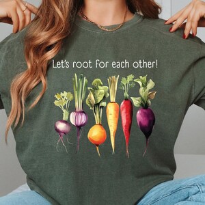 Op de afbeelding: Een groen T-shirt met de tekst "Let's root for each other!" en een afbeelding van kleurrijke groenten, waaronder wortels, rapen en radijzen.