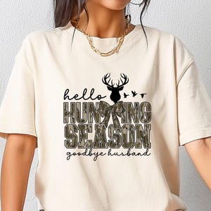 K&ouml;nnte beinhalten: Beiges T-Shirt mit dem Text "hello HUNTING SEASON goodbye husband" in Schwarz und Camouflage-Gr&uuml;n. Das Design beinhaltet eine Hirschsilhouette und V&ouml;gel. Das T-Shirt wird von einer Person getragen.