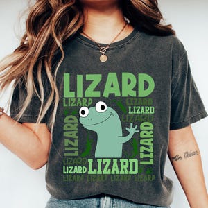 May include: Dark gray t-shirt with a green lizard graphic and the word "LIZARD" repeated. The lizard is light green with large eyes and is waving. The shirt is a casual style.