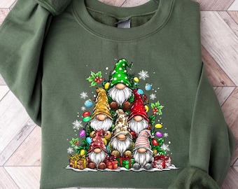 May include: An olive green sweatshirt featuring a Christmas gnome design. The graphic includes multiple gnomes with colorful hats, surrounded by Christmas lights, snowflakes, and wrapped gifts. The sweatshirt has long sleeves and a crew neck.