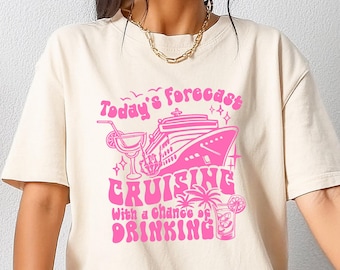 May include: Cream-colored t-shirt featuring a pink graphic that reads "Today's forecast: Cruising with a chance of drinking." The design includes a cruise ship, cocktails, and palm trees. The shirt is a casual, oversized fit.