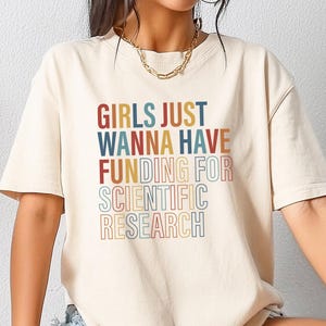 Pode incluir: Camiseta creme com a frase "GIRLS JUST WANNA HAVE FUNDING FOR SCIENTIFIC RESEARCH" em uma fonte retrô colorida. A camiseta tem mangas curtas e um corte folgado.