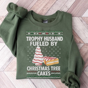 May include: An olive green sweatshirt with the text "TROPHY HUSBAND FUELED BY CHRISTMAS TREE CAKES" in white. The design includes a Christmas tree and a cake. The sweatshirt is folded on a wooden surface.