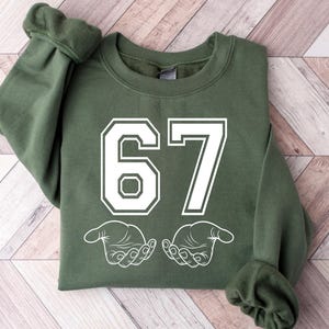May include: An olive green sweatshirt with the number "67" in white block letters. Below the number are two white hands. The sweatshirt is made of a soft material and has cuffed sleeves.