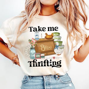 May include: Cream-colored t-shirt with the text "Take me thrifting" in black. The graphic includes a wooden bread box, vintage bowls, a duck, and a jar. The shirt is paired with light blue jeans.