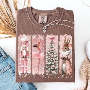 Puede incluir: Camiseta marrón con un diseño festivo que presenta un cascanueces rosa, una bailarina, un árbol de Navidad y un ratón. La camiseta incluye el texto "It's the most wonderful time of the year". También se ven un collar plateado y unos vaqueros azules.