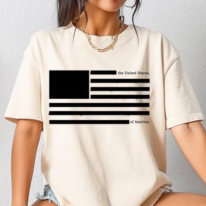 Può includere: T-shirt color crema con grafica in bianco e nero della bandiera americana. Il design della bandiera include un rettangolo nero nell'angolo in alto a sinistra e strisce nere. Il testo "the United States" e "of America" è stampato sopra e sotto la bandiera.
