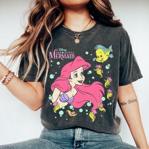Puede incluir: Camiseta gris oscuro con un gr&aacute;fico de Ariel de La Sirenita de Disney. Ariel tiene el pelo rosa y un sujetador de concha p&uacute;rpura. La camiseta tambi&eacute;n incluye ilustraciones de Flounder y caballitos de mar. El texto "Disney The Little Mermaid" est&aacute; encima de Ariel.