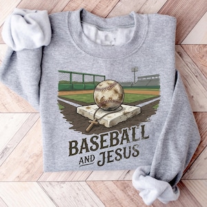 Puede incluir: Una sudadera gris jaspeado de cuello redondo con un gráfico de una pelota de béisbol sobre una base en un estadio. Un collar con una cruz de madera descansa sobre la base. El texto BASEBALL AND JESUS aparece en letras marrón oscuro.