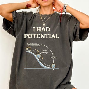 May include: Dark gray t-shirt with the text "I HAD POTENTIAL" above a diagram illustrating potential and kinetic energy. The diagram shows a ball rolling down a curved path, with labels indicating "Potential," "Kinetic Energy," and "NOW."