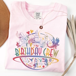 Może przedstawiać: Jasnoróżowa koszulka Comfort Colors z kolorową grafiką "BIRTHDAY CREW". Grafika przedstawia postacie z kreskówek i ikony z popularnych filmów i gier. Złoty naszyjnik jest zarzucony na koszulkę.