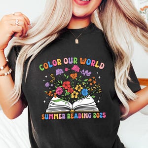 Puede incluir: Camiseta negra con el texto "COLOR OUR WORLD" en colores arcoíris sobre un libro abierto con flores coloridas y el texto "SUMMER READING 2025". La camiseta es de estilo casual y cómodo.